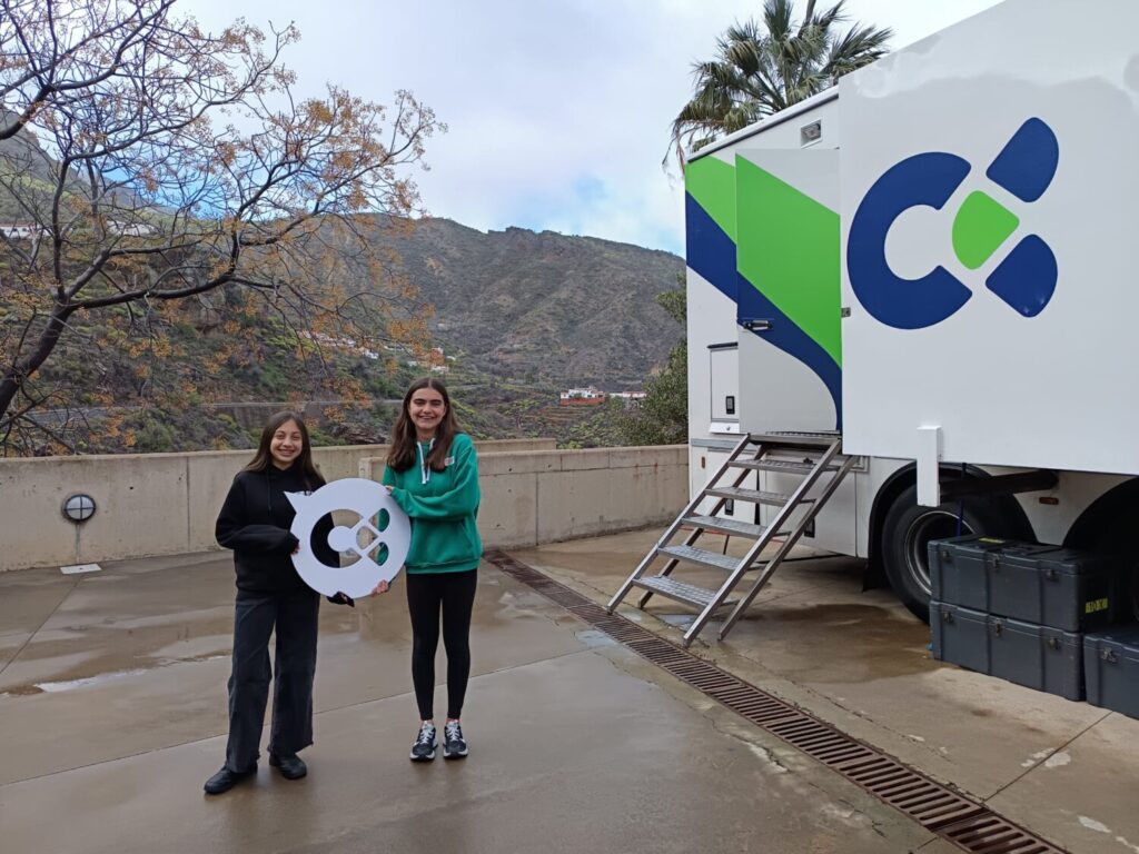 Dos estudiantes del CEO Tejeda junto a la unidad móvil de 'Campus RTVC' sostienen una figura con el logo de Televisión Canaria